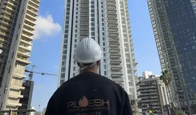 עובד של פילו אש עם מעיל שחור וקסדה לבנה מביט עח שלושה מבנים גבוהים האתר בניה. הפרויקט - איטום מעברי אש